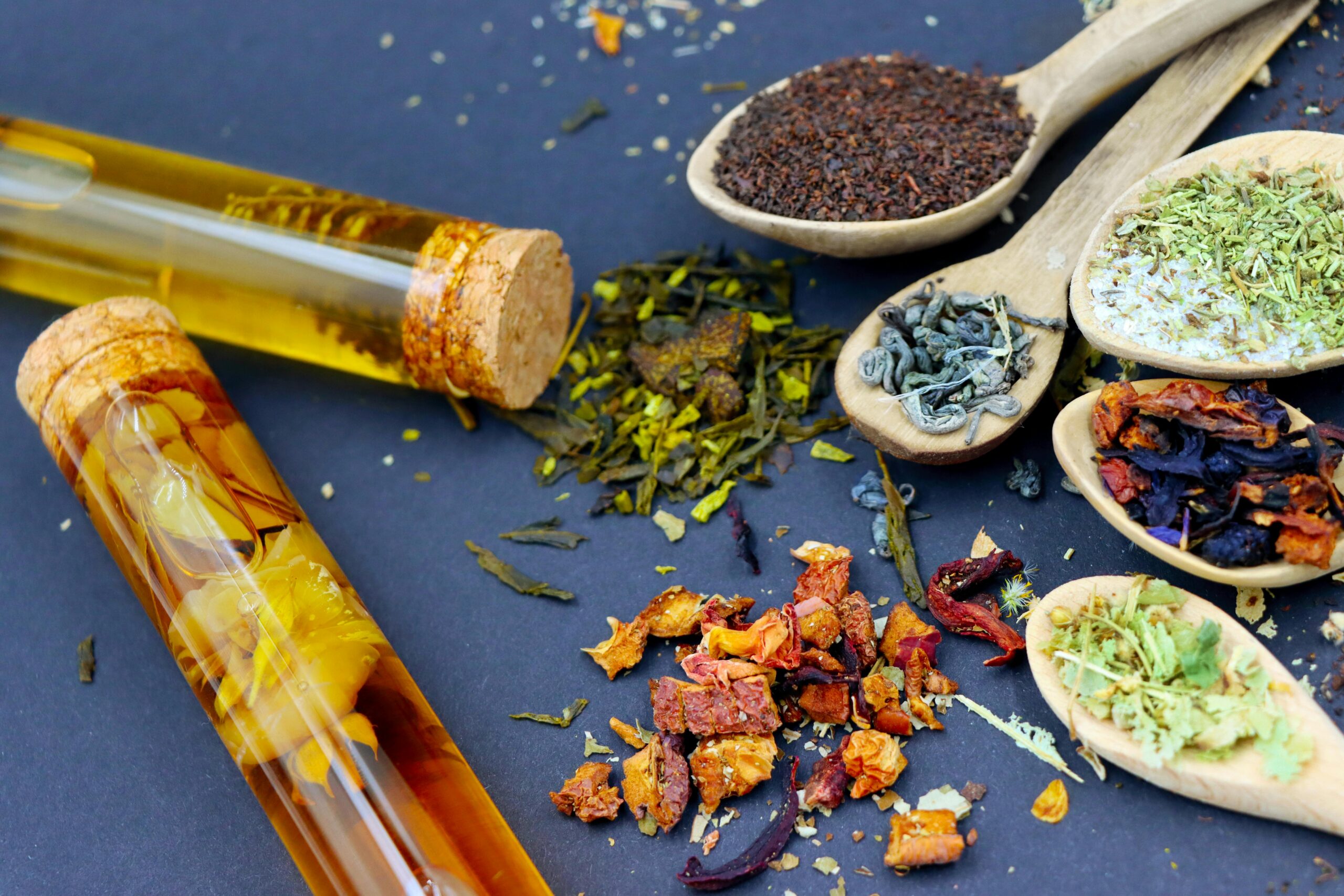 An assortment of dried herbs, tea leaves, and botanicals on wooden spoons with infused herbal oil in glass vials. A perfect representation of natural healing and holistic wellness through herbal remedies.